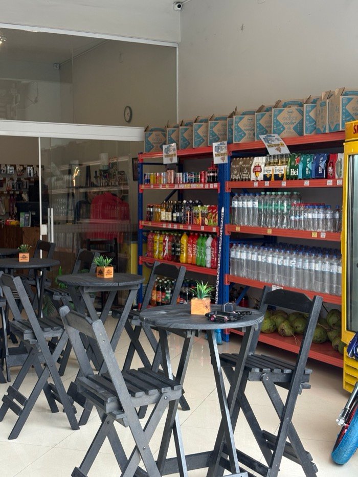 Mesas altas e banquetas de madeira escura na parte interna, com a porta de vidro à esquerda e prateleiras de bebidas ao fundo.
